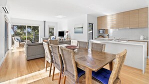 A modern kitchen with a central island, a dining area with a wooden table and wicker chairs, and a living area with a sofa and a fireplace.