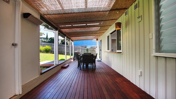 Een overdekt terras met een houten dek, een tafel met stoelen en uitzicht op de achtertuin.