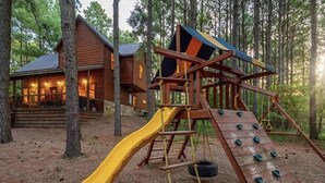 A wooden cabin with a slide and climbing wall in a forest setting. - Bears 'n Biscuits Cabin, Sleeps 14, Hot tub, Playset, Pool Tbl, 4 brms, 3 baths (Broken Bow)