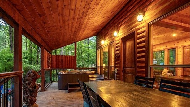 A wooden deck with a dining table and chairs, surrounded by glass doors and windows, overlooking a forested area. - Bears 'n Biscuits Cabin, Sleeps 14, Hot tub, Playset, Pool Tbl, 4 brms, 3 baths (Broken Bow)
