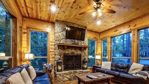 A cozy cabin interior with a stone fireplace, leather sofas, and large windows offering forest views. - Bears 'n Biscuits Cabin, Sleeps 14, Hot tub, Playset, Pool Tbl, 4 brms, 3 baths (Broken Bow)