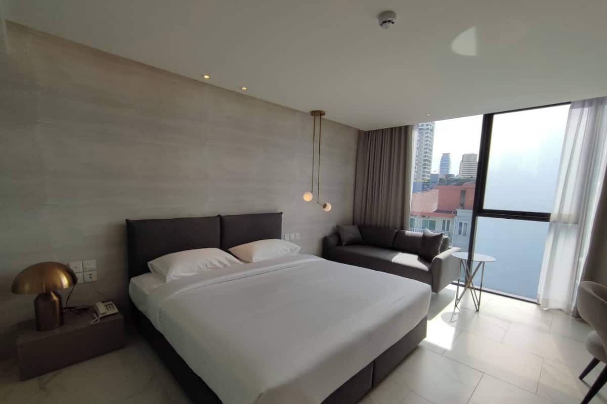 Quarto de hotel moderno com uma cama grande, um sofá, uma mesinha e vista para a cidade através de grandes janelas.