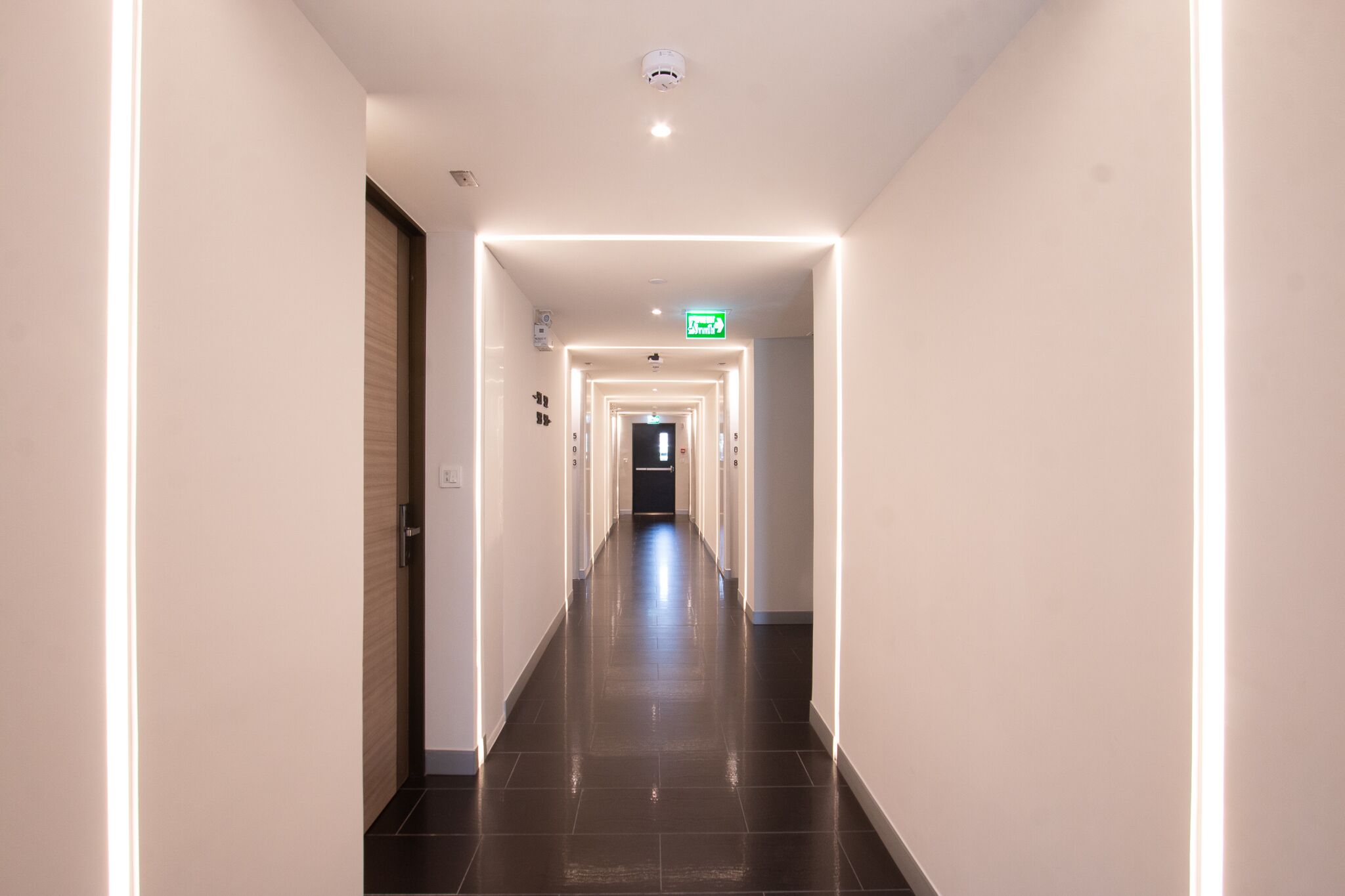 A modern hotel hallway with a long, straight corridor, illuminated by recessed lighting, and featuring a central exit sign.