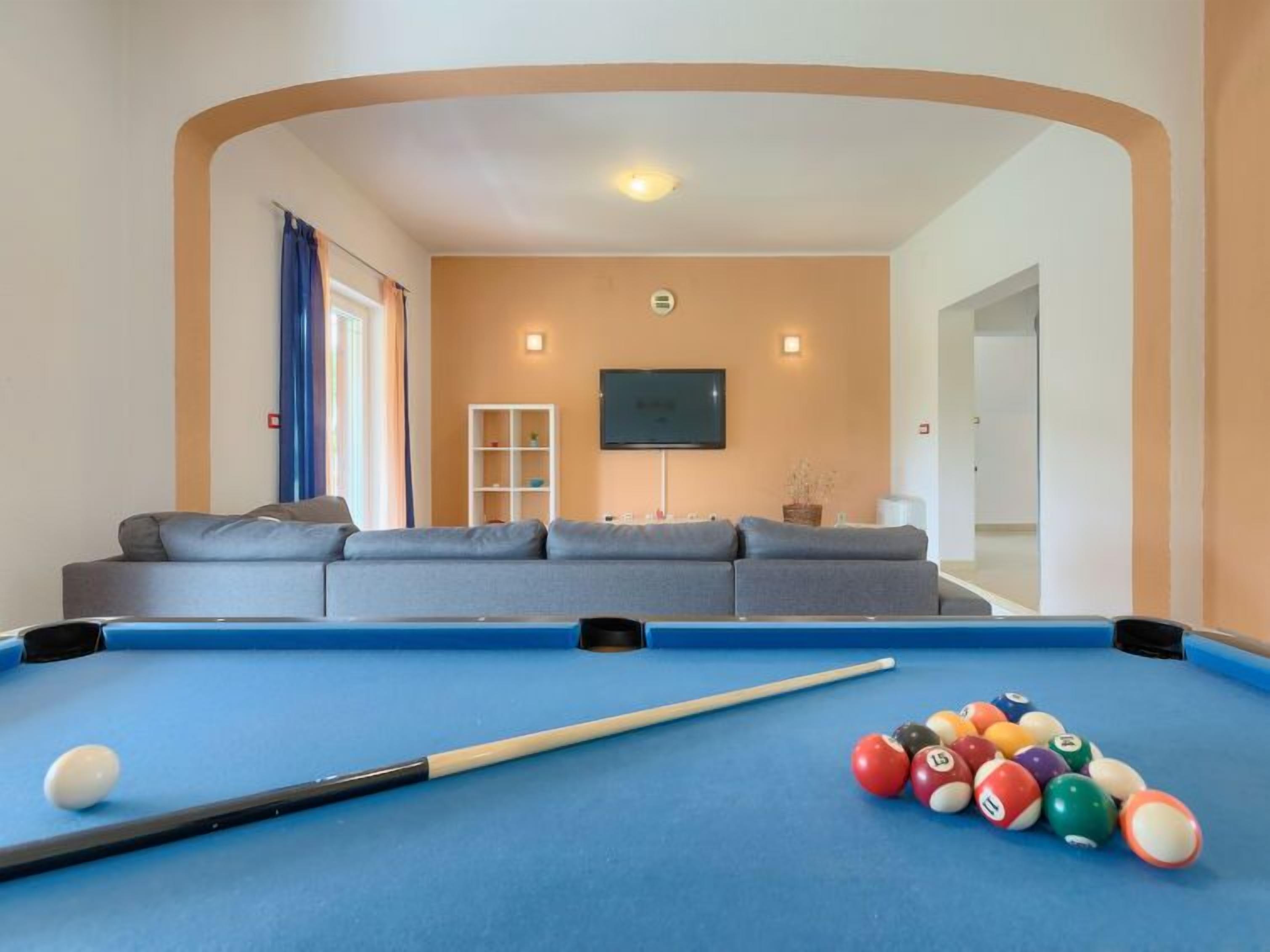 Une table de billard avec un queue et des billes, un canapé et un téléviseur dans une pièce dotée d’un plafond incurvé et de murs de couleur beige.