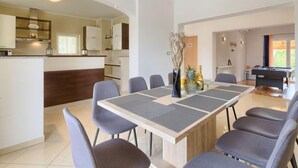 Un espace repas moderne avec une table rectangulaire et six chaises, une cuisine aux meubles blancs et, au fond, une table de billard.