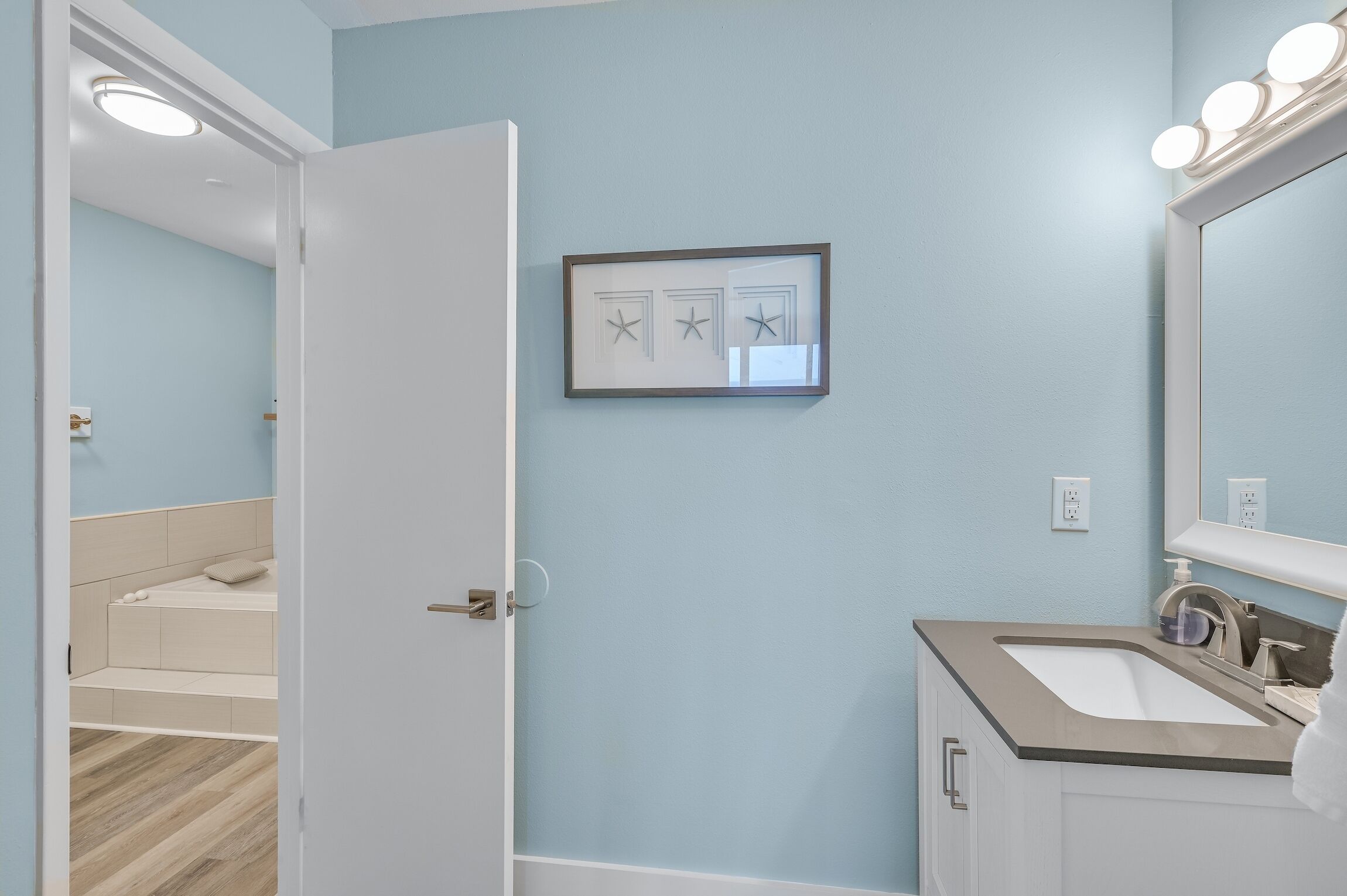 A bathroom with a light blue wall, a framed picture, a sink with a mirror, and a door leading to a bedroom.