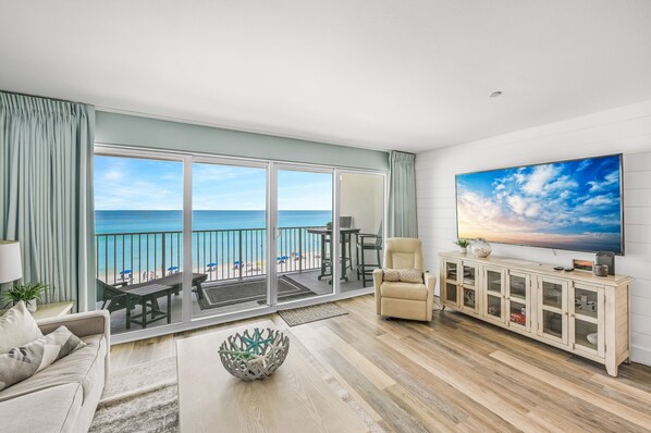 Amplia sala de estar con un gran ventanal con vista a la playa, un sofá cómodo, un televisor y un mueble de madera.