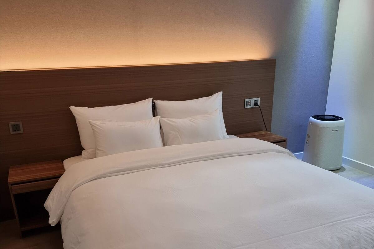 Un lit bien fait, avec du linge de lit blanc, une tête de lit en bois et une table de chevet surmontée d’une lampe.
