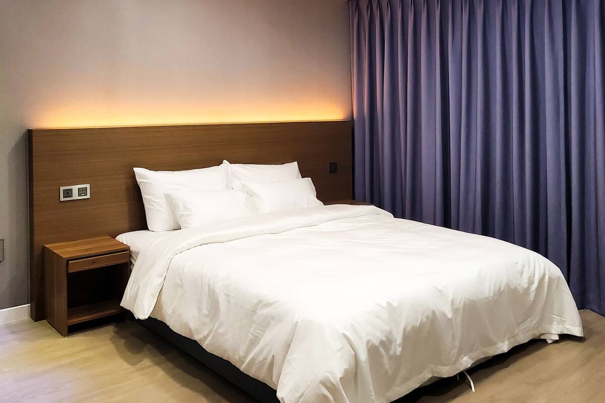 Un lit bien fait, avec du linge de lit blanc, une tête de lit en bois et une table de chevet dotée d’une prise électrique.