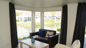 A living room with a sofa, a coffee table, and a chair. There is a window with a view of a house and a body of water. - Comfortable Chalet Near Brunssummerheide (Brunssum)