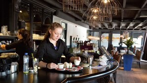 Een bar met een toonbank, barkrukken en iemand die een drankje bereidt.