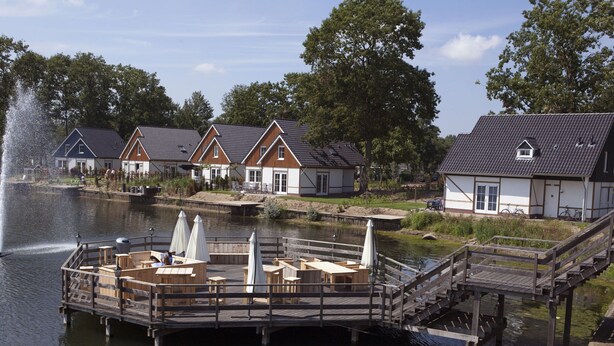 Een houten steiger aan een meer met zitplaatsen en een fontein, omringd door traditionele huizen met schuine daken.