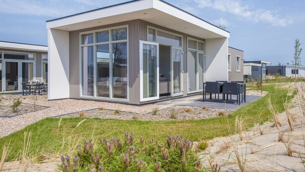 Un bâtiment moderne, de plain-pied, doté de grandes fenêtres en verre et de portes coulissantes, d’un patio avec des meubles d’extérieur, et d’un paysage sablonneux à la végétation clairsemée.