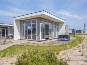 Un bâtiment moderne, de plain-pied, doté de grandes fenêtres en verre et de portes coulissantes, d’un patio avec des meubles d’extérieur, et d’un paysage sablonneux à la végétation clairsemée.