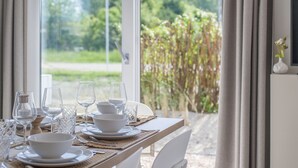 Une table à manger est dressée avec de l’assiette blanche, des bols et des verres à vin, et est placée près d’une fenêtre donnant sur un paysage verdoyant et sur un drapeau à l’extérieur.