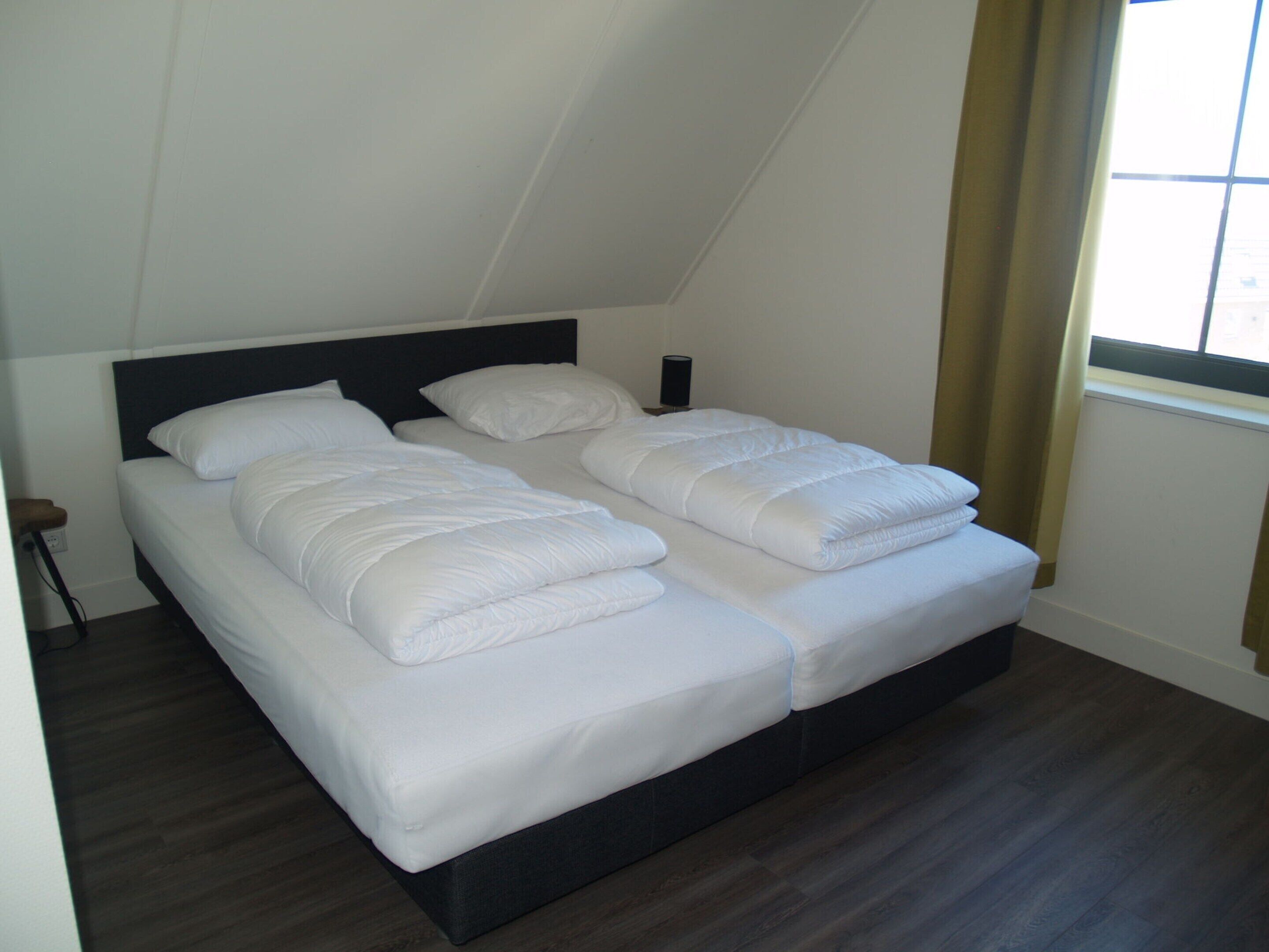 Een tweepersoonsbed met een donker hoofdeinde in een kamer met een schuine plafond en een raam met gordijnen.