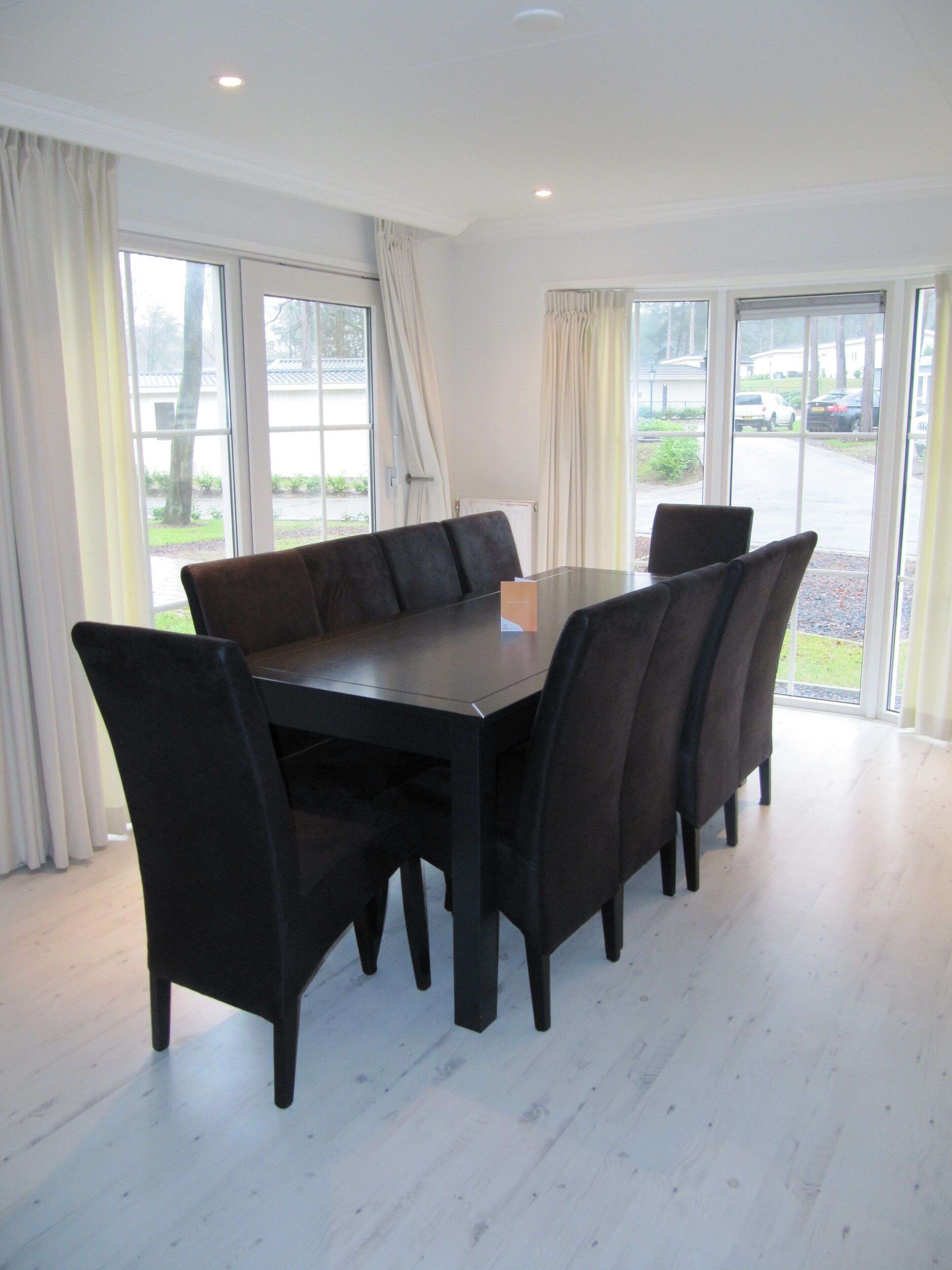 A dining area with a rectangular table and six chairs, large windows with curtains, and a view of a parking area outside.