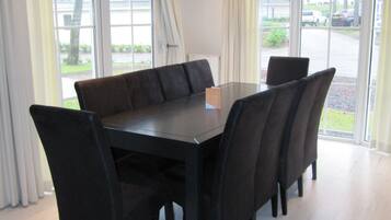 A dining area with a rectangular table and six chairs, large windows with curtains, and a view of a parking area outside.