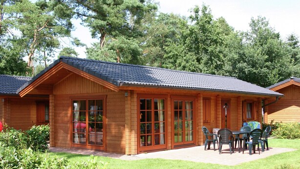 A wooden cabin with a tiled roof, large glass doors, and a patio with outdoor furniture. - Wooden Chalet Located in the Achterhoek (Lochem)