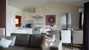 A modern living room with a grey sofa, a dining area with a white table and chairs, a kitchen with white cabinets and a gas stove, and a purple wall with a wreath. - Wooden Chalet Located in the Achterhoek (Lochem)