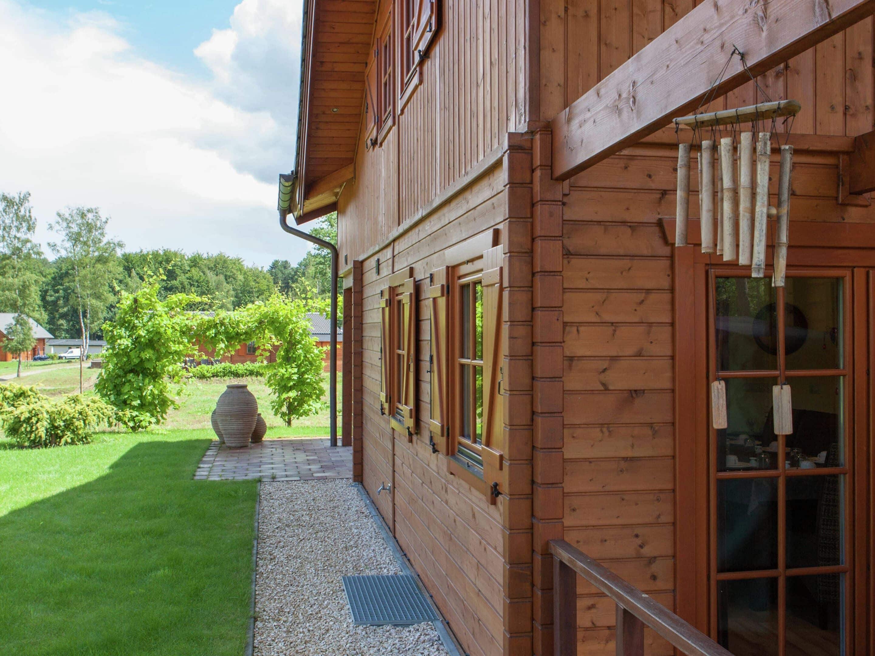Ein Holzhaus mit überdachter Veranda, einem großen Fenster und Blick auf Garten und Bäume.