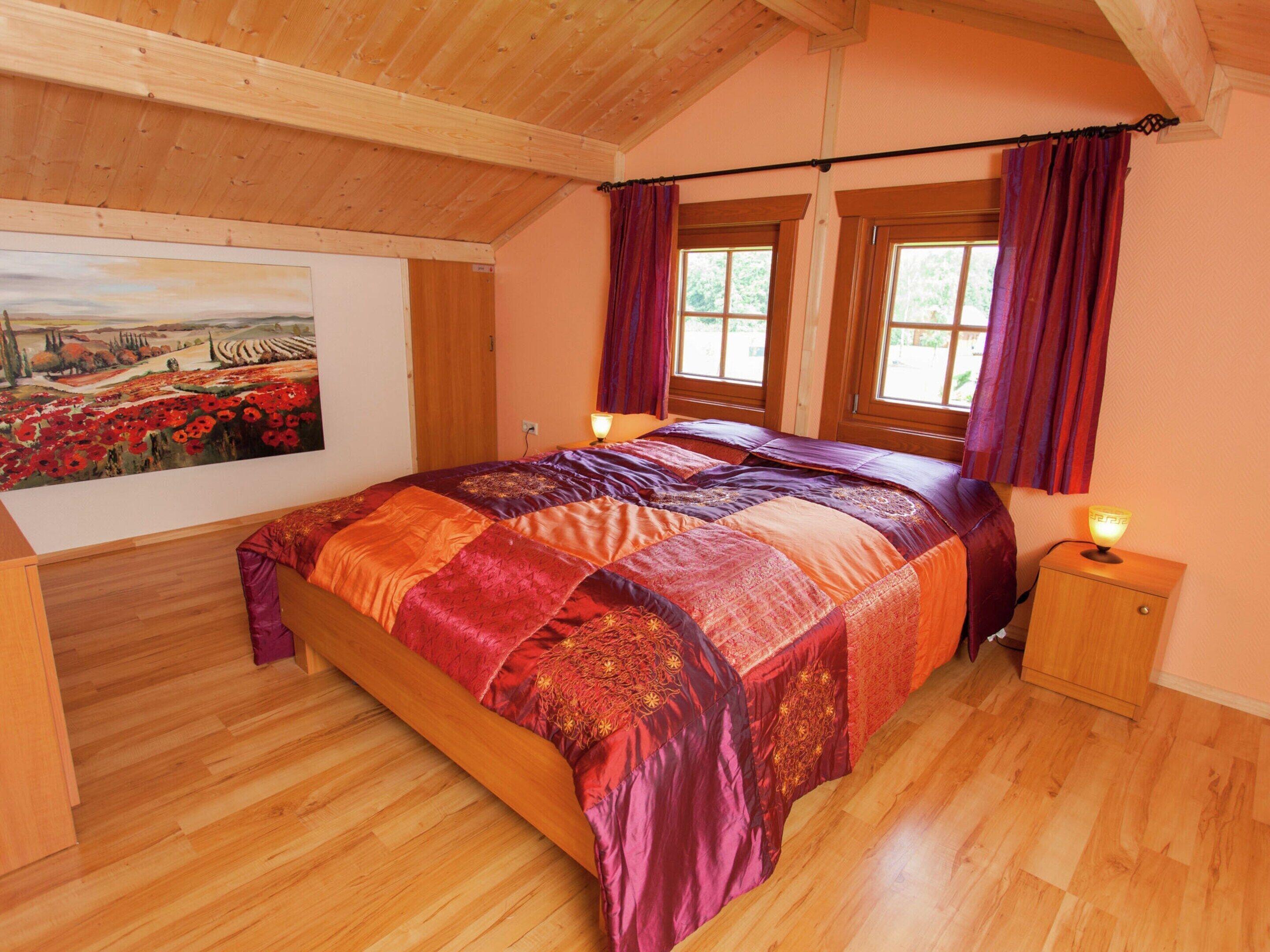 Ein Schlafzimmer mit einem großen Bett, Holzböden, einem Bild an der Wand und zwei mit Vorhängen versehenen Fenstern.