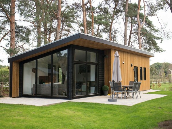 A modern, single-story house with large glass windows, a patio with outdoor furniture, and a wooden exterior.