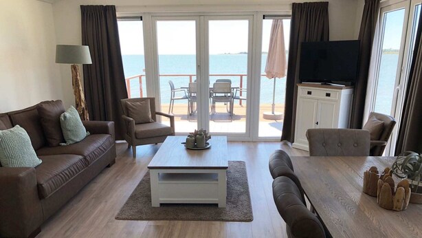Uma sala de estar espaçosa, com sofá, poltrona e mesa de jantar. Uma televisão está fixada na parede, e há vista para o mar através das portas de vidro.