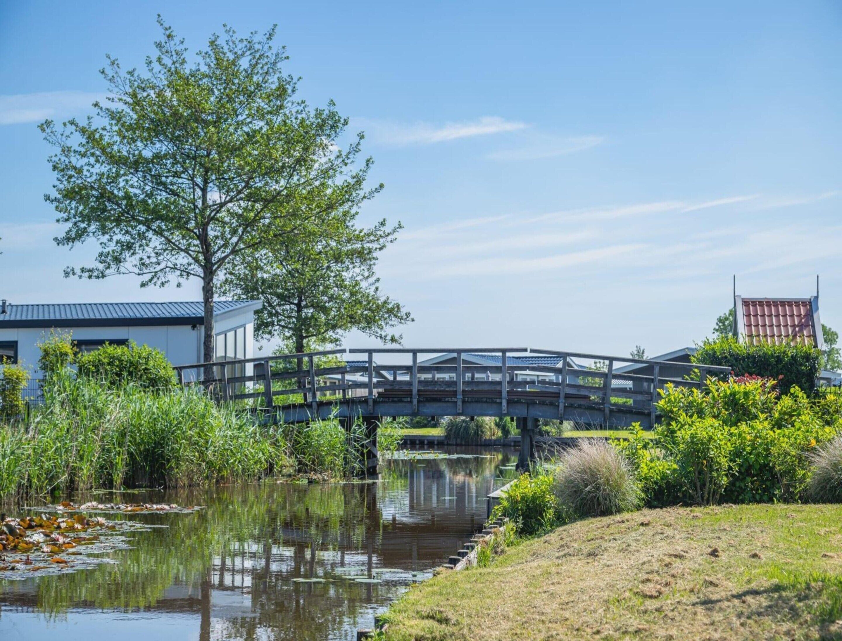 A bridge over a calm water body with a grassy bank, surrounded by greenery and buildings.