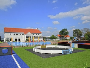 A swimming pool area with a circular water feature, a covered seating area with umbrellas, and a building with a red roof in the background. - Luxury Chalet With a Dishwasher (Nunspeet)