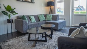 A modern living room with a grey sectional sofa, a round coffee table, a floor lamp, a potted plant, and a window with a view of a house. - Dutch Family Geataway in West-graftdijk (West-Graftdijk)