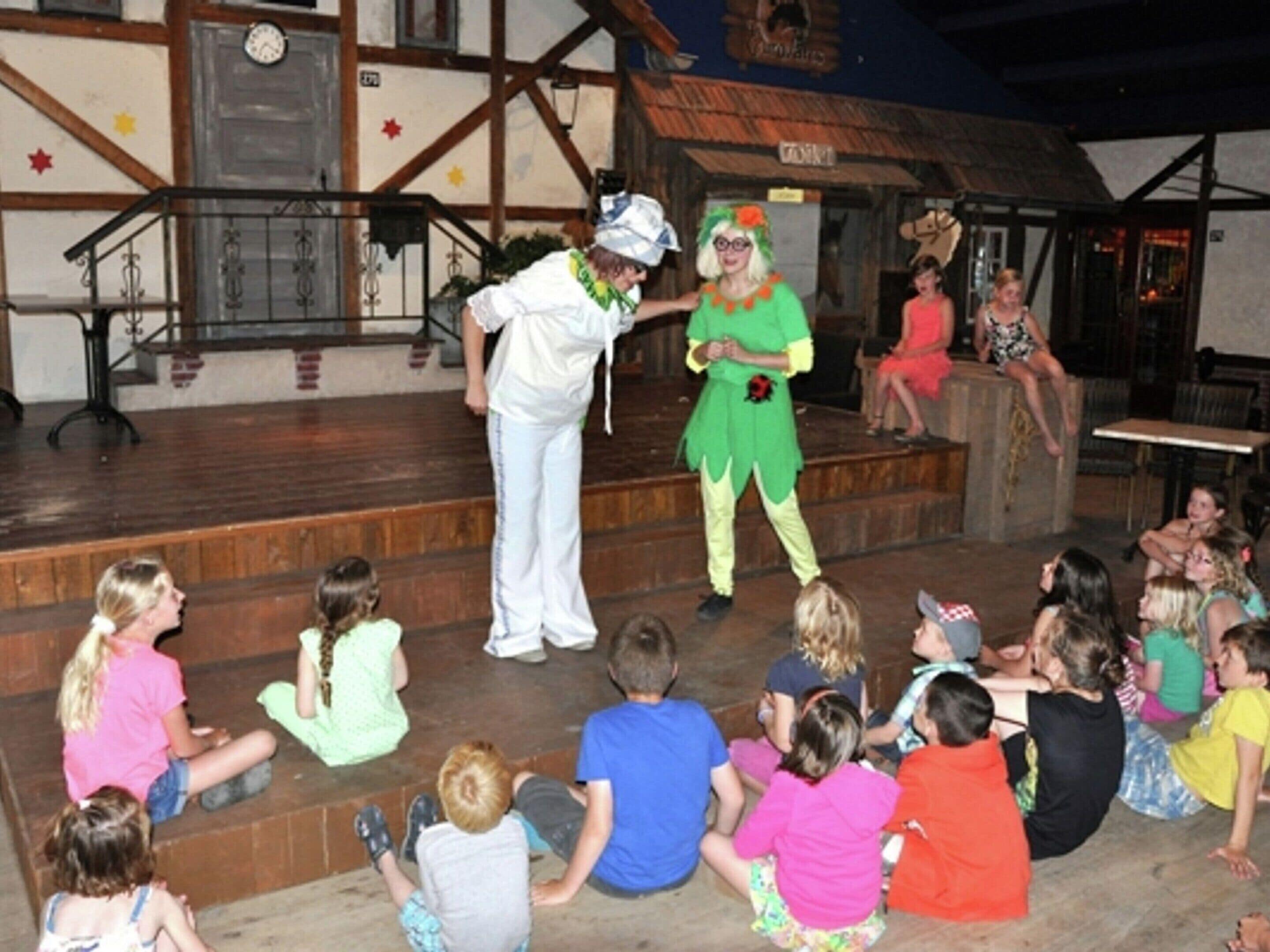 A stage with two performers in costumes, children seated in front, and a rustic building in the background.