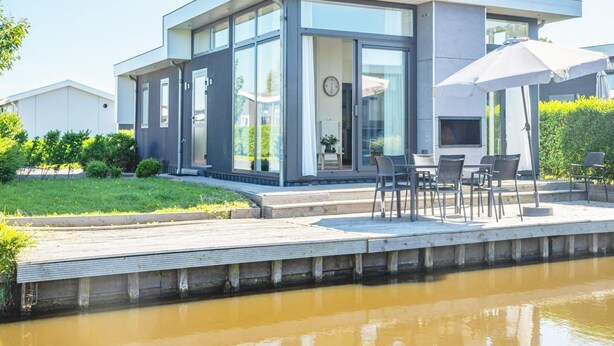Ein modernes Haus mit Glasfront, einer Terrasse mit Gartenmöbeln und einem Fluss im Vordergrund.