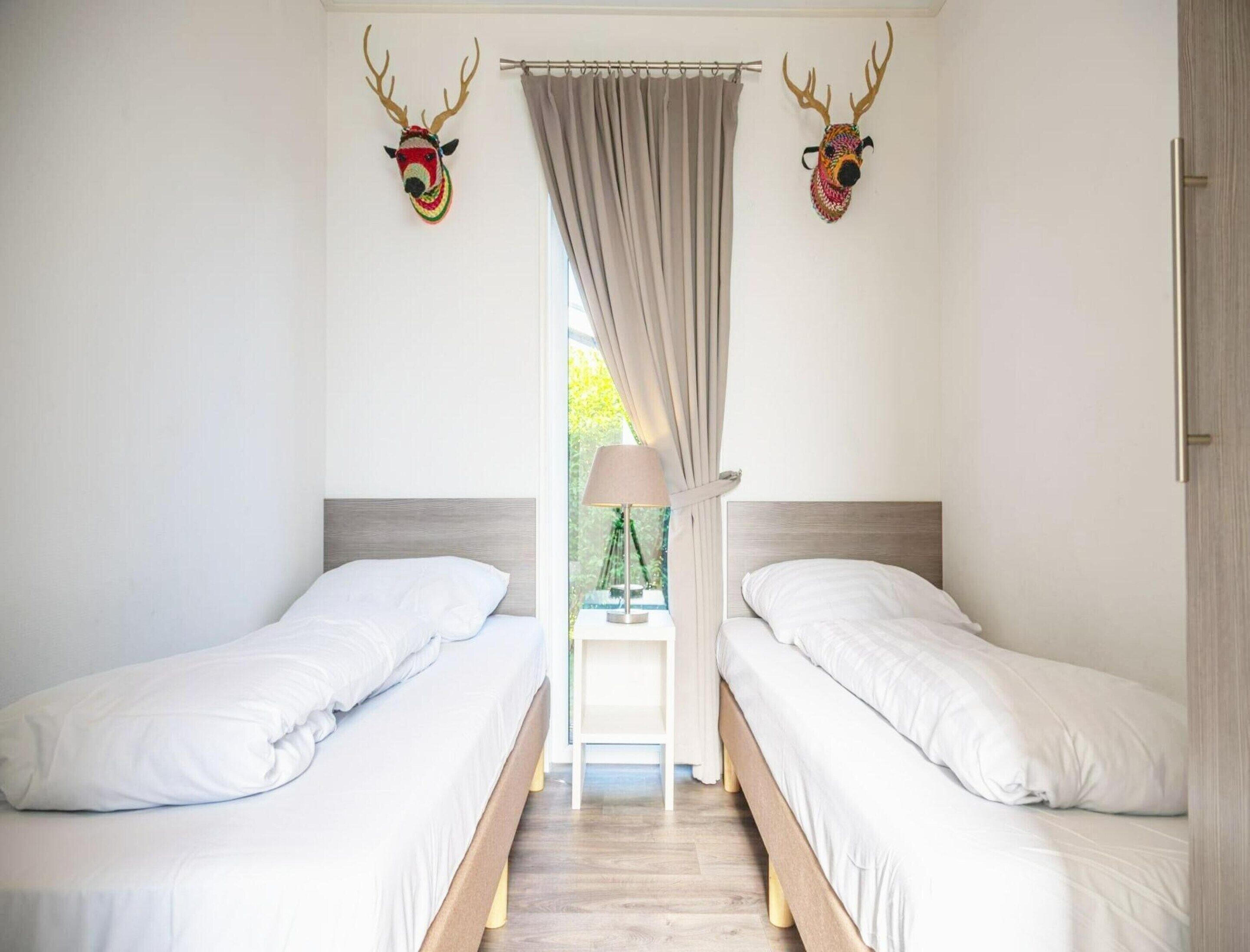Une chambre avec deux lits simples, une fenêtre avec des rideaux et deux têtes d’animaux décoratives accrochées aux murs.