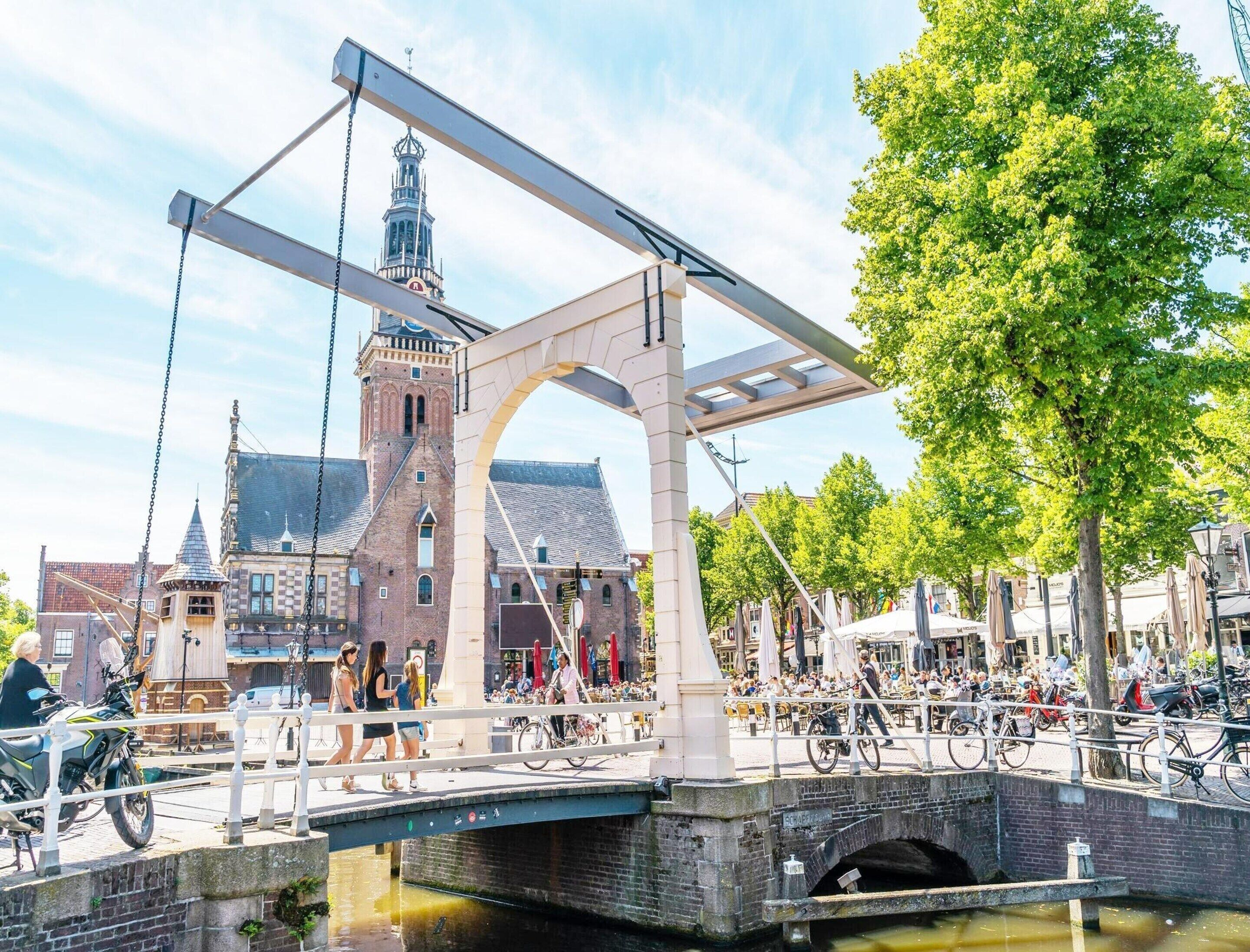 A bridge with a metal arch structure over a canal, a historic building with a tower in the background, and a tree-lined street with bicycles and pedestrians.