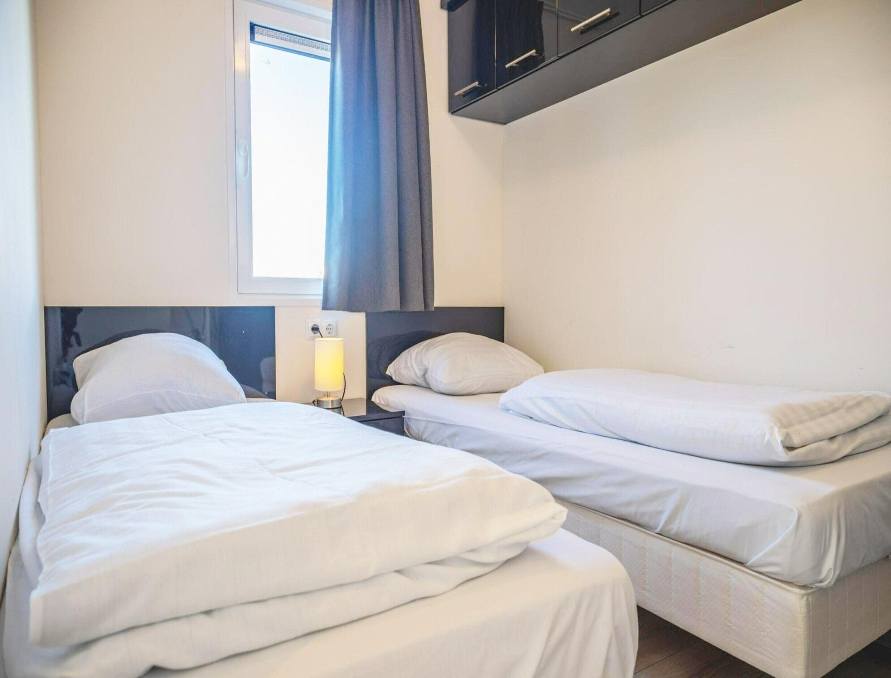 A hotel room with two beds, a window with curtains, a bedside lamp, and a dark-colored wardrobe.