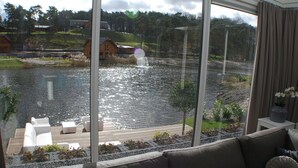 A modern living room with a large window overlooking a lake, a wooden deck, and a view of houses across the water. - Modern Holiday Home Near Brunssummerheide (Brunssum)