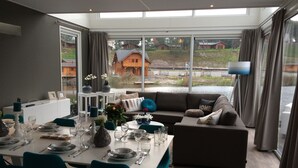 A modern dining area with a table set for four, a sofa, and a view of a lake and houses. - Modern Holiday Home Near Brunssummerheide (Brunssum)