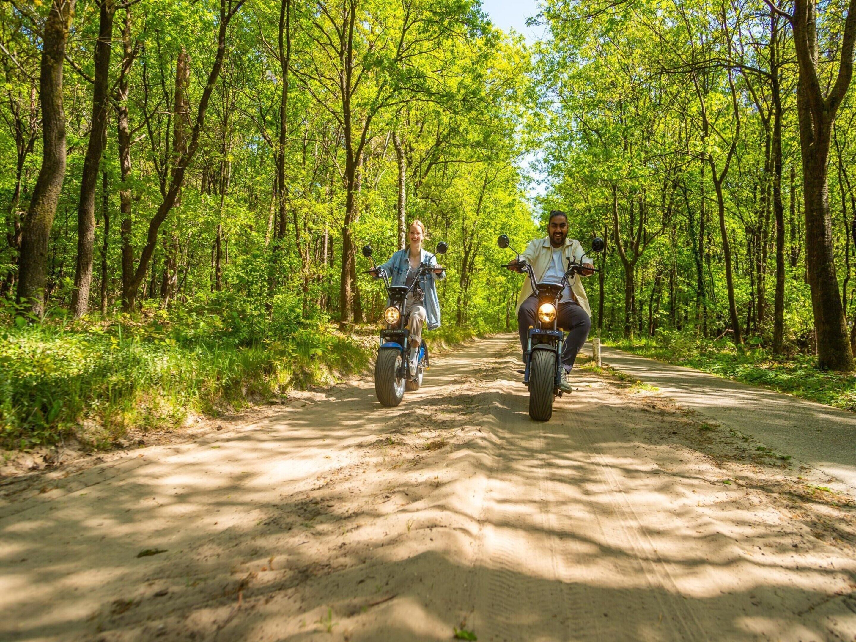 Two people riding motorcycles on a dirt path surrounded by trees.
