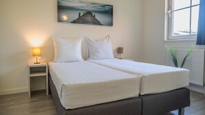 Uma cama de casal com roupa de cama branca, uma mesa de cabeceira com abajur, uma janela com vista e um quadro na parede.