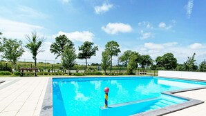 Una piscina rectangular con un borde empedrado, un banco y árboles al fondo.