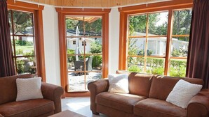 A living room with a brown leather sofa, a wooden coffee table, and large windows with a view of a house and greenery outside. - Wooden Chalet With Terrace and Microwave (Brunssum)