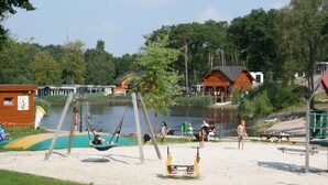 A lakeside park with a swing set, picnic tables, and a sandy area. - Wooden Chalet With Terrace and Microwave (Brunssum)