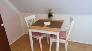 Een klein zitehoekje met een houten tafel en twee witte stoelen met rood-wit geblokte kussens. Op de tafel staat een pot met gele bloemen.