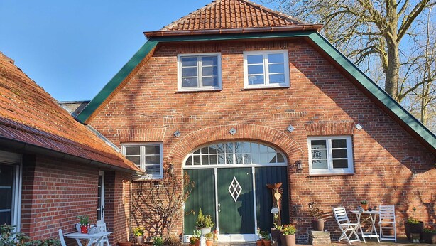 Een bakstenen huis met een groene deur, twee ramen en een kleine tuin met potplanten en tuinmeubilair.