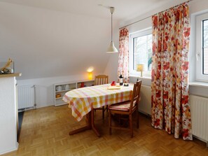 Un comedor con una mesa de madera, sillas, mantel a cuadros, una lámpara colgante y cortinas con estampado floral.