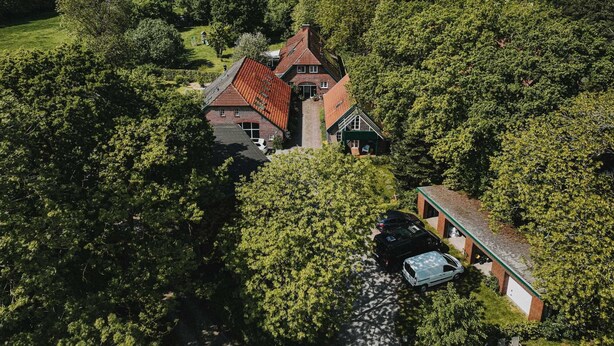 A large, two-story house with a red roof, surrounded by dense greenery and a small driveway with two cars.