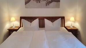 A neatly made bed with white linens and two pillows, flanked by two bedside tables with lamps, in a room with a wooden headboard and a painting of hands on the wall. - Deichhof Dangast, WH Löwenzahn - Holiday apartment Löwenzahn (Varel)