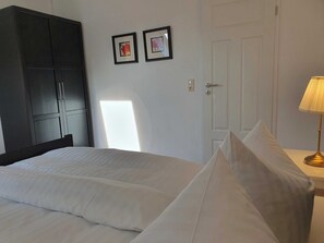 A neatly made bed with white linens, a dark wooden wardrobe, two framed pictures on the wall, and a bedside lamp.
