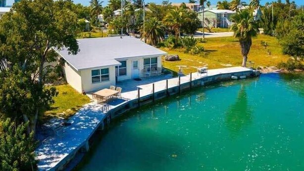 A modern house with a dock extending into a clear blue water body, surrounded by lush greenery and palm trees. - Fish Camp | Old Florida canalfront getaway (Key West)